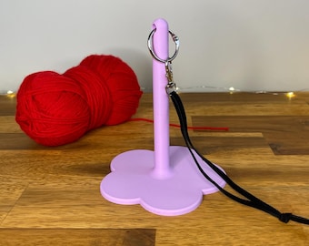 May include: A light purple yarn holder with a flower-shaped base and a silver keyring. A black lanyard is attached to the keyring. Two balls of red yarn are in the background. The text "3D PRINTING FILES | STL" is at the bottom.