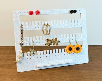 May include: A white felt earring display stand with various earrings, including gold hoops, floral designs, and sunflower-shaped pairs. Red buttons and black studs are also displayed. The text "3D PRINTING FILES | STL" is visible at the bottom of the image.