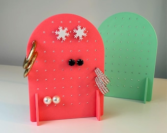 May include: Two earring display stands, one coral and one green, with multiple holes for earrings. The coral stand has snowflake accents and holds various earrings, including hoops and studs. The bottom of the image reads "3D PRINTING FILES | STL".
