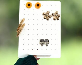 May include: A white earring display board with various earrings. The earrings include gold hoops, sunflower studs, gold flower studs, and skull studs. The text "3D PRINTING FILES | STL" is at the bottom of the image.
