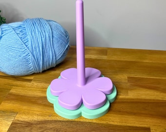 May include: A light purple and green yarn holder in the shape of a flower with a tall, vertical post. A large ball of light blue yarn sits to the left of the holder. The text "3D PRINTING FILES | STL" is at the bottom.