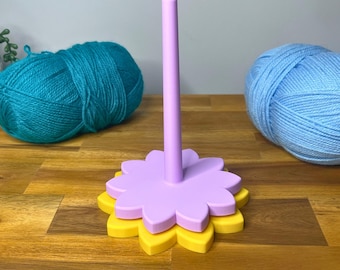 May include: A purple and yellow yarn holder in the shape of a flower. The yarn holder has a central post and a base with a flower design. Two balls of yarn, one teal and one light blue, are in the background. The text "3D PRINTING FILES | STL" is at the bottom.