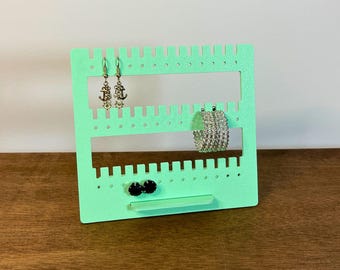 May include: A mint green earring display stand with multiple slots and holes for hanging jewellery. The stand holds various earrings, including silver anchor drop earrings, a silver bracelet, and black stud earrings. The text "3D PRINTING FILES | STL" is at the bottom.