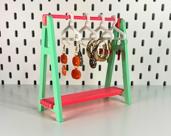 May include: A miniature jewellery display stand in mint green and pink. The stand features a pink shelf and top bar, with white hangers holding earrings. These include pumpkin-shaped and gold hoop styles. The text "3D PRINTING FILES | STL" is at the bottom.