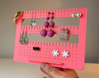 May include: A vibrant pink earring display stand, showcasing a variety of earrings and a bracelet. The stand features slots and holes for hanging jewellery. The text "3D PRINTING FILES | STL" is visible at the bottom.