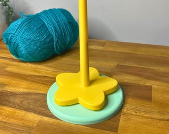 May include: A yellow flower-shaped yarn holder with a mint green base and a tall yellow center post. A ball of teal yarn sits to the left. The text "3D PRINTING FILES | STL" is at the bottom.