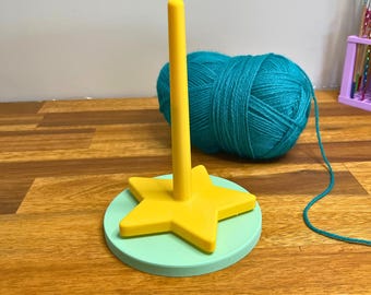May include: A yellow star-shaped yarn holder with a teal base and a vertical post. A ball of teal yarn sits behind the holder. The base is on a wooden surface. The text "3D PRINTING FILES | STL" is at the bottom of the image.