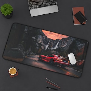 May include: A red Dodge Viper sports car driving on a winding road through a mountainous landscape with waterfalls in the background. The car is driving towards the viewer and the landscape is a mix of green trees and rocky cliffs.