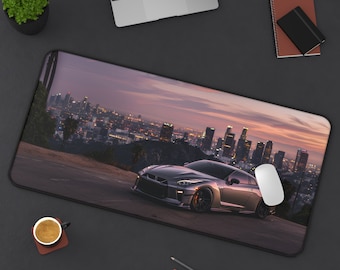 May include: A gray sports car with a city skyline in the background. The car is on a road with a mouse pad in the foreground. The city skyline is lit up at night.