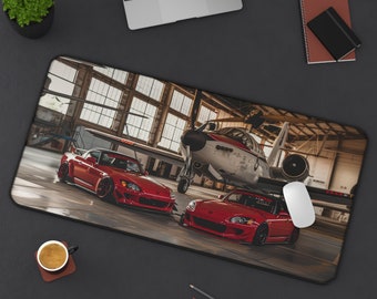 May include: A red Honda S2000 sports car and a red Honda Civic Si sports car are parked in a hangar with a red, white, and blue airplane in the background. The cars are both facing the camera and have their headlights on.