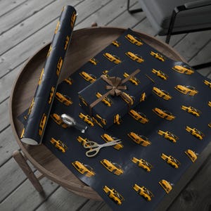 May include: A roll of dark blue wrapping paper and a rectangular gift box wrapped in the same paper, both featuring a repeating pattern of yellow sports cars. A pair of silver scissors and a tape dispenser are placed on the flat sheet of paper.