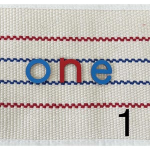 Op de afbeelding: Blauwe en rode lettertegels die het woord "one" spellen op een witte stoffen achtergrond met rode en blauwe strepen. Het cijfer "1" staat in het zwart onder het woord.