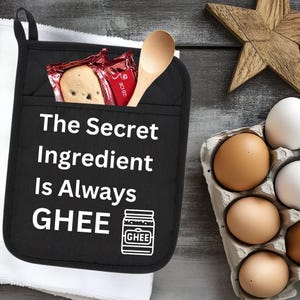 Puede incluir: Manopla de horno negra con texto blanco que dice "The Secret Ingredient Is Always GHEE" y una ilustración blanca de un tarro de ghee. La manopla está sobre una toalla blanca con una cuchara de madera y un paquete de galletas.