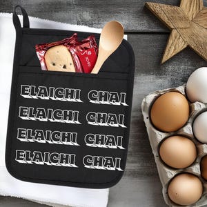 May include: Black oven mitt with the text "ELAICHI CHAI" in white. A wooden spoon and packaged cookies are in the mitt's pocket. A carton of eggs and a wooden star are in the background.
