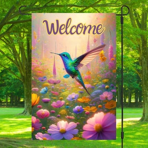 May include: A garden flag with a hummingbird flying over a field of colorful flowers. The flag has the word "Welcome" written in purple letters.