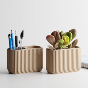 May include: Two beige, ribbed desk organizers. One holds pens and pencils, the other a succulent plant. The organizers are rectangular with rounded corners. A small notebook is visible in the background.