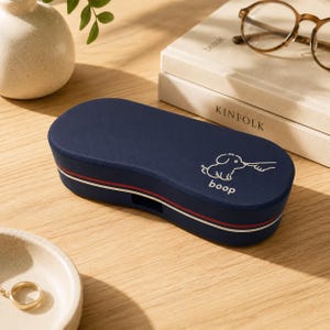 May include: A navy blue, oval-shaped hard-shell glasses case featuring a white line-art graphic of a finger touching a puppy's nose with the text "boop" printed below. The case has a thin red and white horizontal stripe along the side.