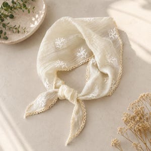 May include: A cream-colored triangular headscarf made of a lightweight, textured fabric. The scarf features delicate white floral embroidery scattered across the surface and is finished with a decorative scalloped crochet trim along the edges.
