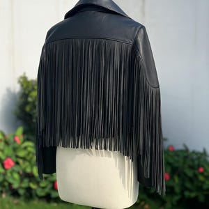May include: Black leather jacket with fringe detail. The jacket has a classic collar and long fringe along the back. The jacket is displayed on a mannequin in front of a green background.