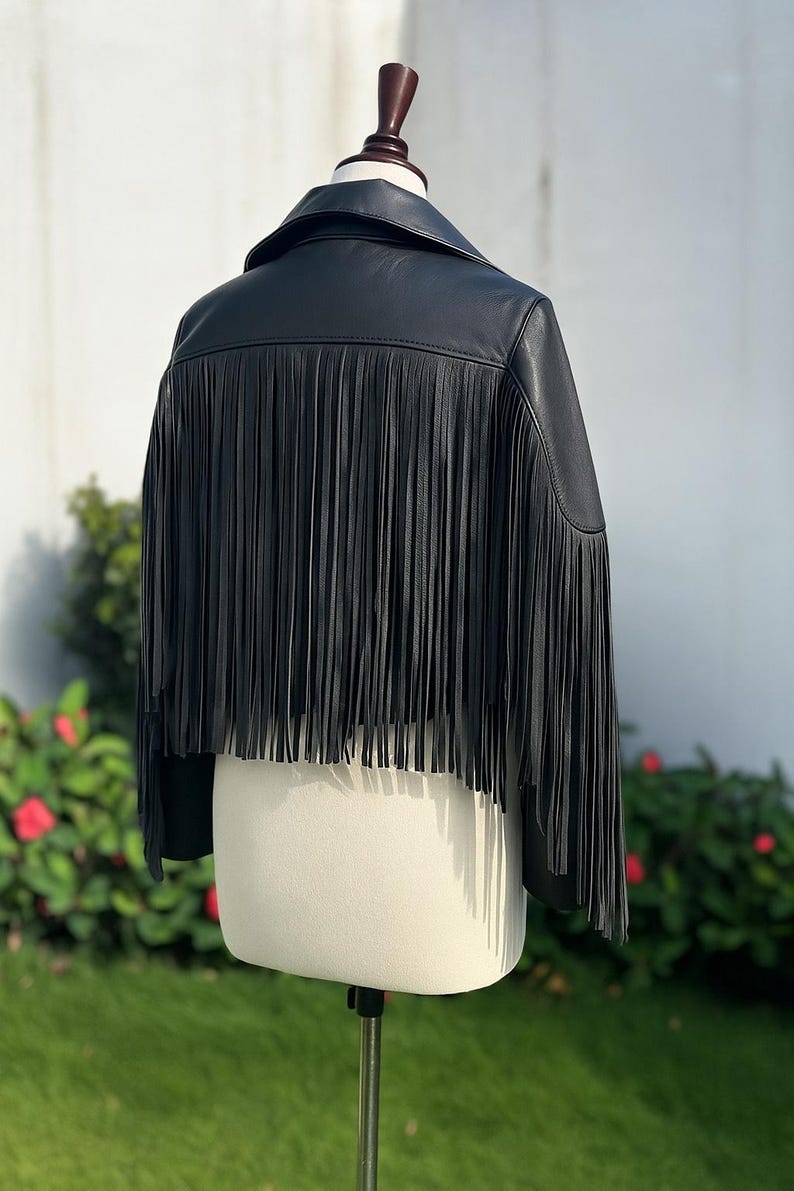 May include: Black leather jacket with fringe detail. The jacket has a classic collar and long fringe along the back. The jacket is displayed on a mannequin in front of a green background.