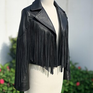 May include: A black leather jacket with long fringe detailing. The jacket has a classic collar and is displayed on a mannequin. The fringe cascades down the front and sleeves, adding a stylish, edgy look. This jacket is a statement piece.