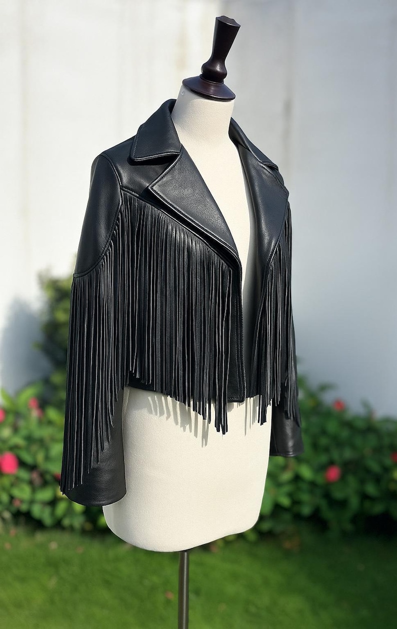 May include: A black leather jacket with long fringe detailing. The jacket has a classic collar and is displayed on a mannequin. The fringe cascades down the front and sleeves, adding a stylish, edgy look. This jacket is a statement piece.