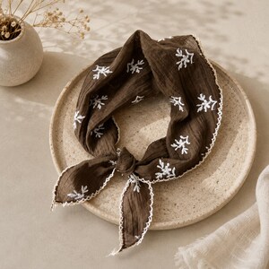 May include: A dark brown square scarf made of crinkled fabric featuring white embroidered floral sprigs scattered across the surface. The edges of the scarf are finished with a delicate white scalloped lace trim, and it is tied in a simple knot.
