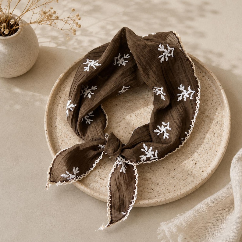 May include: A dark brown square scarf made of crinkled fabric featuring white embroidered floral sprigs scattered across the surface. The edges of the scarf are finished with a delicate white scalloped lace trim, and it is tied in a simple knot.