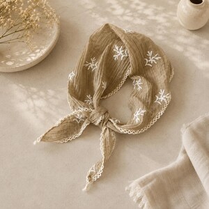 May include: A tan triangular headscarf made of crinkled fabric featuring white embroidered floral sprigs and a delicate white scalloped lace trim along the edges. The scarf is tied in a knot at the front, displaying a textured, lightweight appearance.