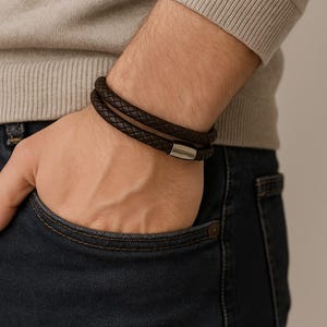 May include: A dark brown leather bracelet with a braided design. The bracelet wraps around the wrist twice and features a silver-colored magnetic clasp. The person is wearing a light gray sweater and dark blue jeans.