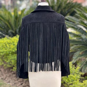 May include: Black suede jacket with long fringe detail. The jacket has a classic collar and is displayed on a mannequin. The fringe hangs from the back and sleeves, adding a stylish, bohemian flair to the garment.