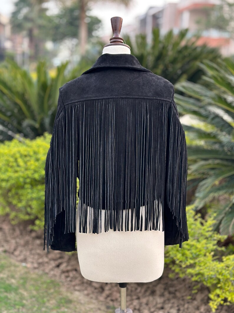 May include: Black suede jacket with long fringe detail. The jacket has a classic collar and is displayed on a mannequin. The fringe hangs from the back and sleeves, adding a stylish, bohemian flair to the garment.