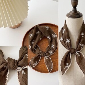 May include: A dark brown square scarf featuring a white embroidered floral pattern and a scalloped white crochet edge. The fabric has a crinkled texture, and the scarf is tied in a knot, displaying the intricate stitching and decorative border.