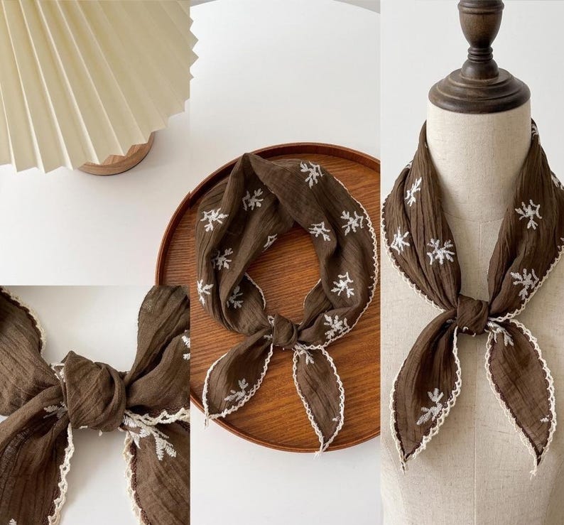 May include: A dark brown square scarf featuring a white embroidered floral pattern and a scalloped white crochet edge. The fabric has a crinkled texture, and the scarf is tied in a knot, displaying the intricate stitching and decorative border.