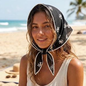 May include: A black headscarf featuring a white embroidered pattern of small coral-like shapes. The triangular fabric is tied under the chin and finished with a delicate white scalloped lace trim along all edges, creating a textured appearance.