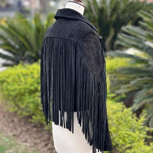 May include: A black suede jacket with long fringe detailing on the back. The jacket is displayed on a mannequin, showcasing its design and texture. The fringe adds a bohemian flair to the garment.
