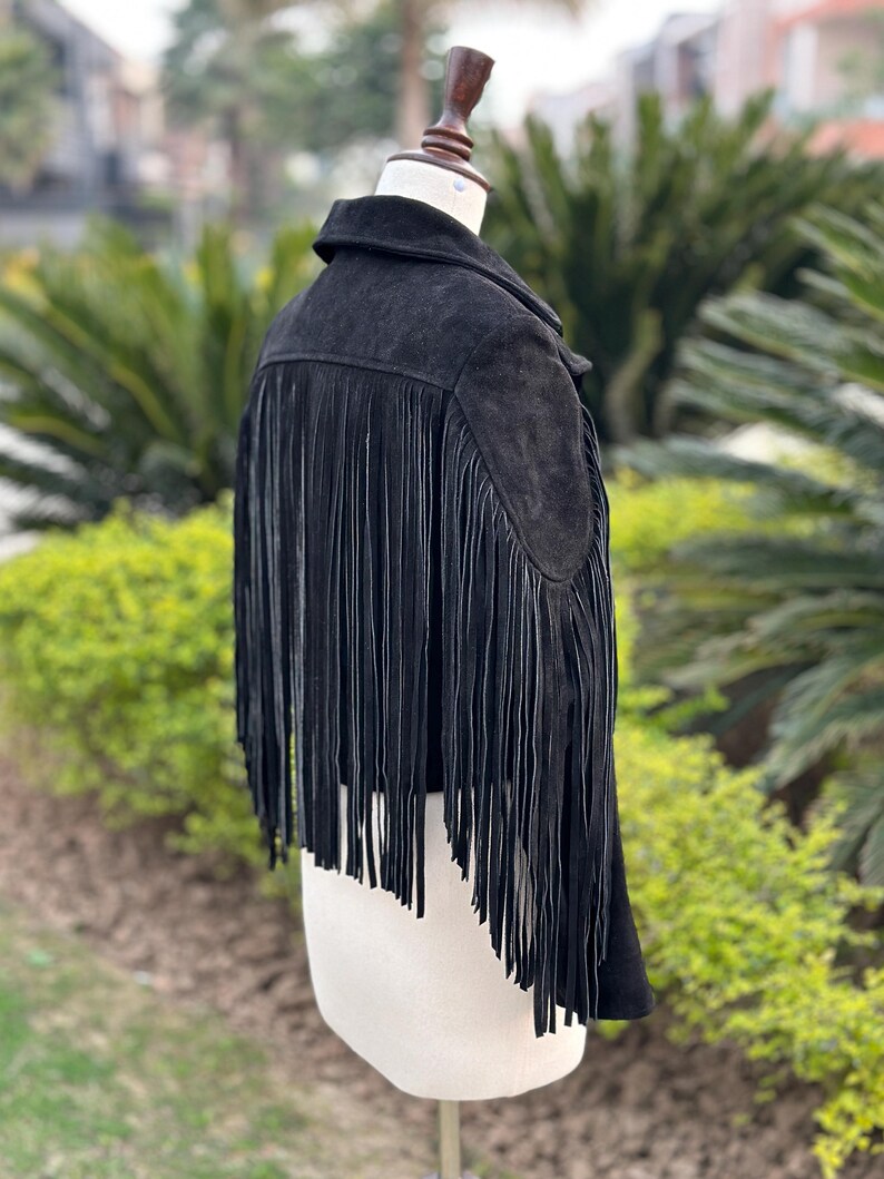May include: A black suede jacket with long fringe detailing on the back. The jacket is displayed on a mannequin, showcasing its design and texture. The fringe adds a bohemian flair to the garment.