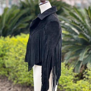 May include: Black suede jacket with long fringe detailing. The jacket features a classic collar and a cropped silhouette. The fringe cascades down the sleeves and across the back, adding a bohemian flair. The jacket is displayed on a mannequin.