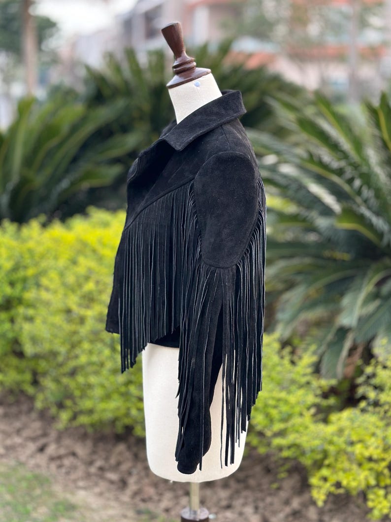 May include: Black suede jacket with long fringe detailing. The jacket features a classic collar and a cropped silhouette. The fringe cascades down the sleeves and across the back, adding a bohemian flair. The jacket is displayed on a mannequin.