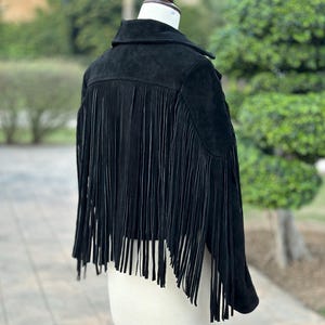 May include: A black suede jacket with long fringe detailing on the back. The jacket is displayed on a mannequin, showcasing its collar and tailored fit. The fringe adds a stylish, textured element to the garment.