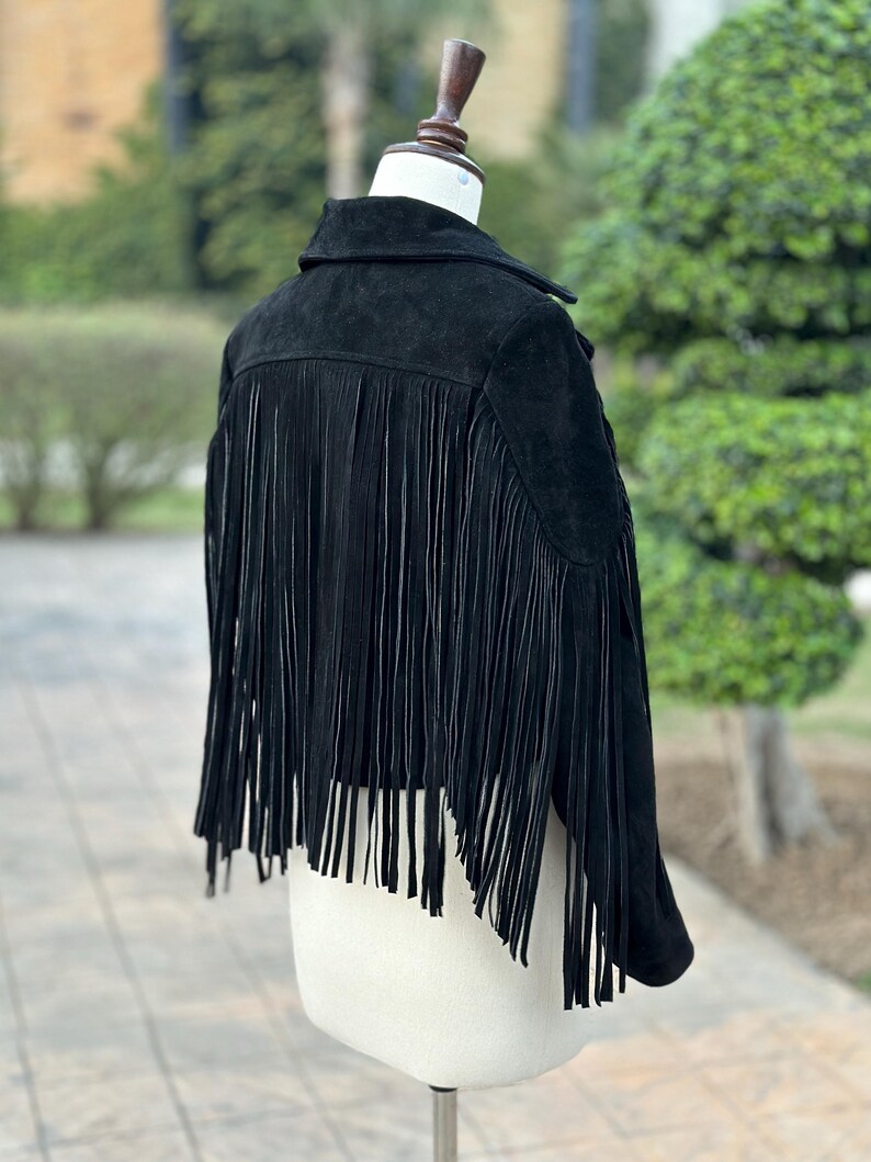 May include: A black suede jacket with long fringe detailing on the back. The jacket is displayed on a mannequin, showcasing its collar and tailored fit. The fringe adds a stylish, textured element to the garment.