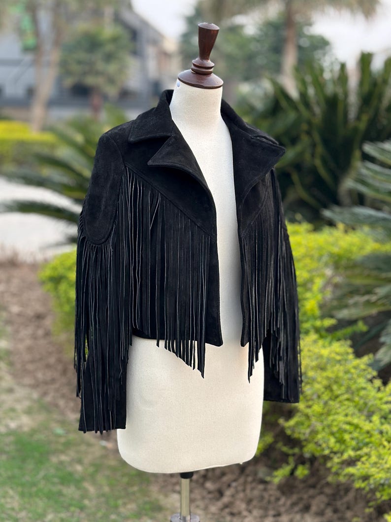 May include: A black suede jacket with fringe detailing. The jacket features a classic collar and a cropped silhouette. The fringe is long and hangs from the sleeves and the hemline, adding a bohemian flair. The jacket is displayed on a mannequin.
