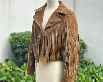 May include: A brown suede jacket with fringe detailing. The jacket is displayed on a mannequin and features a classic collar and a cropped length. The fringe is along the sleeves and the front of the jacket.