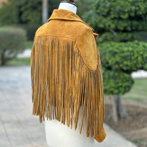 May include: A brown suede jacket with fringe detailing. The jacket has a collar and long sleeves. The fringe is on the bottom of the jacket and the sleeves.