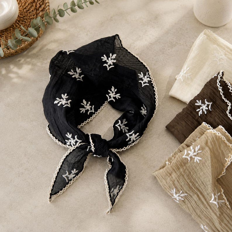 May include: A black square scarf featuring white embroidered floral sprigs and a scalloped white lace edge. The fabric has a crinkled texture. Additional folded scarves in cream, dark brown, and tan are visible nearby, each with matching embroidery.