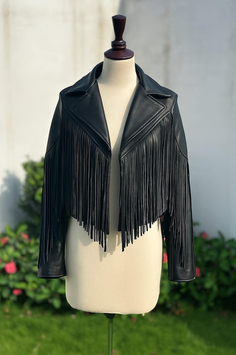May include: A black leather jacket with fringe detailing. The jacket has a classic collar, long sleeves, and a cropped length. The fringe hangs from the chest and sleeves, adding a stylish, bohemian flair. The jacket is displayed on a mannequin.