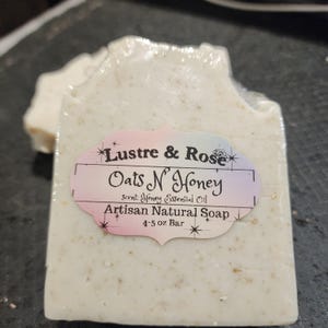May include: A bar of artisan natural soap, wrapped in clear plastic. The soap is a light beige color with visible flecks. A label reads "Lustre & Rose Oats N' Honey" with "Scent: Honey Essential Oil" and "4-5 oz Bar".