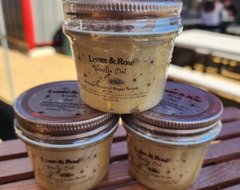 May include: Three glass jars of Vanilla Oat Artisan Natural Sugar Scrub. Each jar has a brown lid and a label with the text "Lustre & Rose Vanilla Oat". The jars are stacked on a wooden surface.