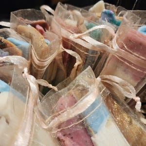 May include: Close-up of several small, colorful soaps in sheer pink organza bags. The soaps are various shapes and colors, including blue, pink, and brown. Each bag is tied with a matching pink ribbon, creating a delicate and gift-ready presentation.