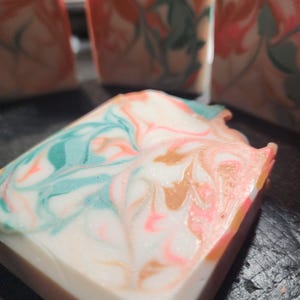 May include: Close-up of a handmade soap bar with a swirled design. The soap features a white base with swirls of teal, pink, and gold. Additional soap bars are visible in the background, showcasing a similar design. The soap has a rectangular shape.